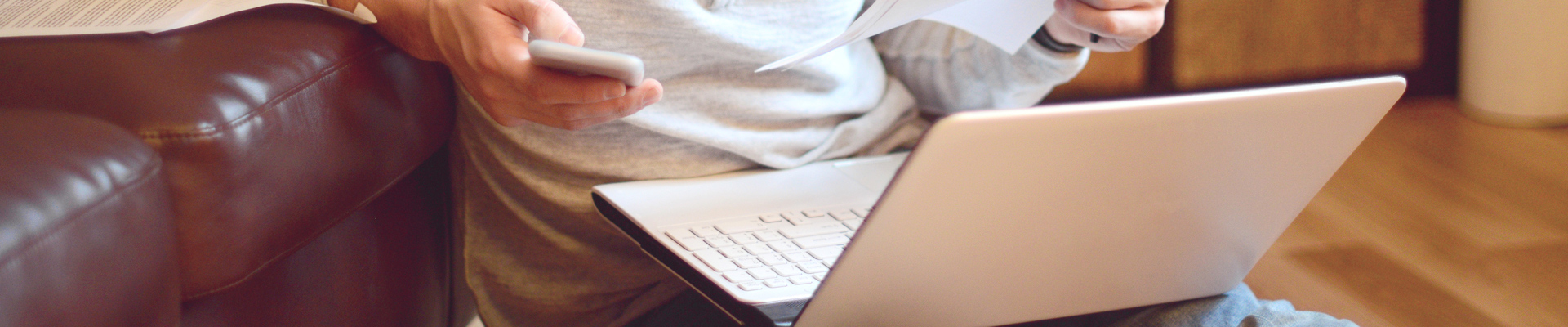 A man sits in front of a laptop holding a phone in one hand and a document in the other